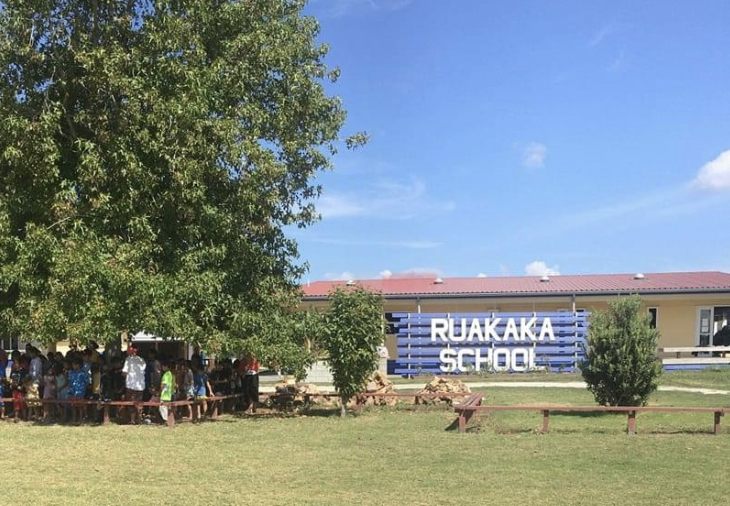 A picture of a school with its big sign. The sign reads Ruakaka School. Benches to sit on at the front. Sunny day. Children grouped in the shade under a big tree.